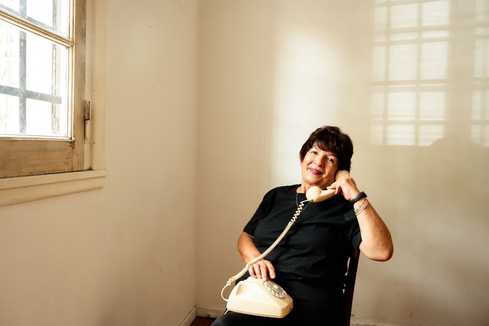 Une dame âgée souriante, combiné filaire à la main, assise près d'une fenêtre baignée de lumière chaude